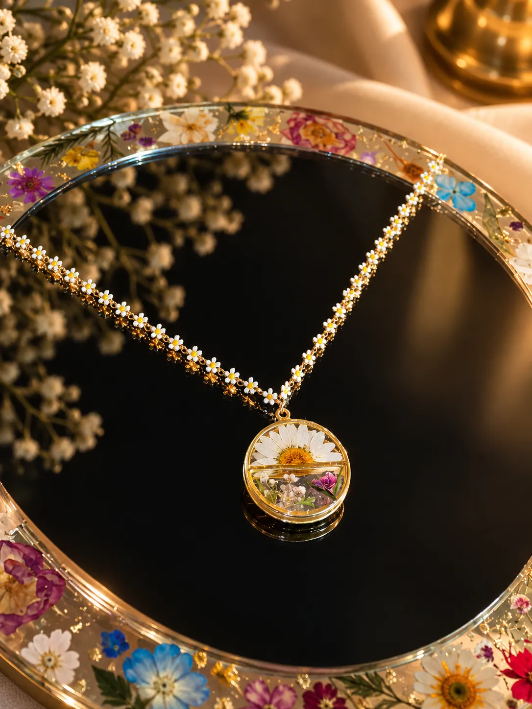 A gold pendant holding a pressed daisy in clear resin, resting on a floral resin tray